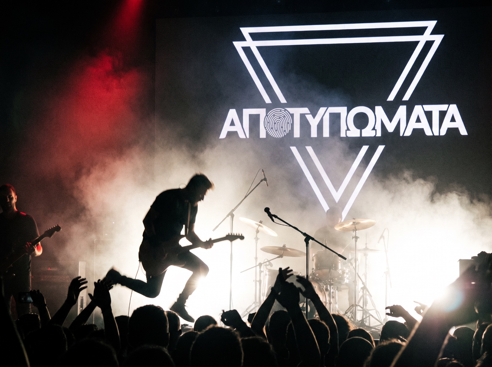 Apotypomata mid-set: a guitarist jumping in silhouette against blinding backlight and smoke, the band emblem lit on the backdrop screen, raised hands in the front rows